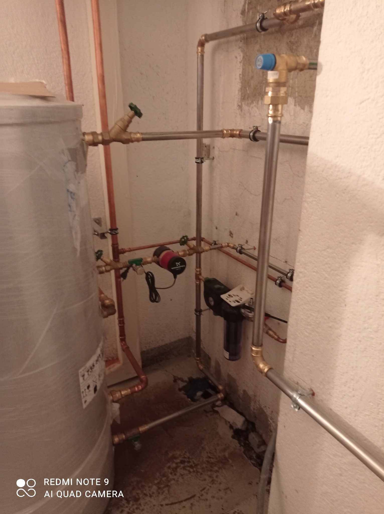 Instalacja grzewcza z szarym zbiornikiem, rurami miedzianymi i stalowymi, pompą obiegową oraz filtrem, zamontowana w pomieszczeniu technicznym z surowymi ścianami.