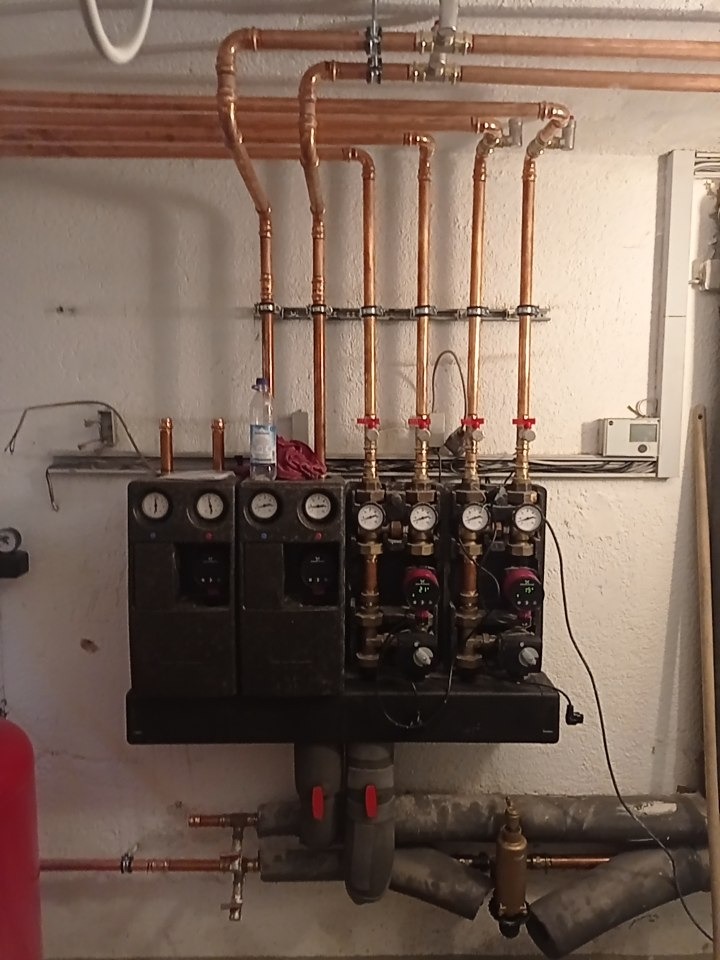 Rozdzielacz ogrzewania podłogowego z rurami miedzianymi, manometrami i zaworami, widoczne butelka wody i ścierka.