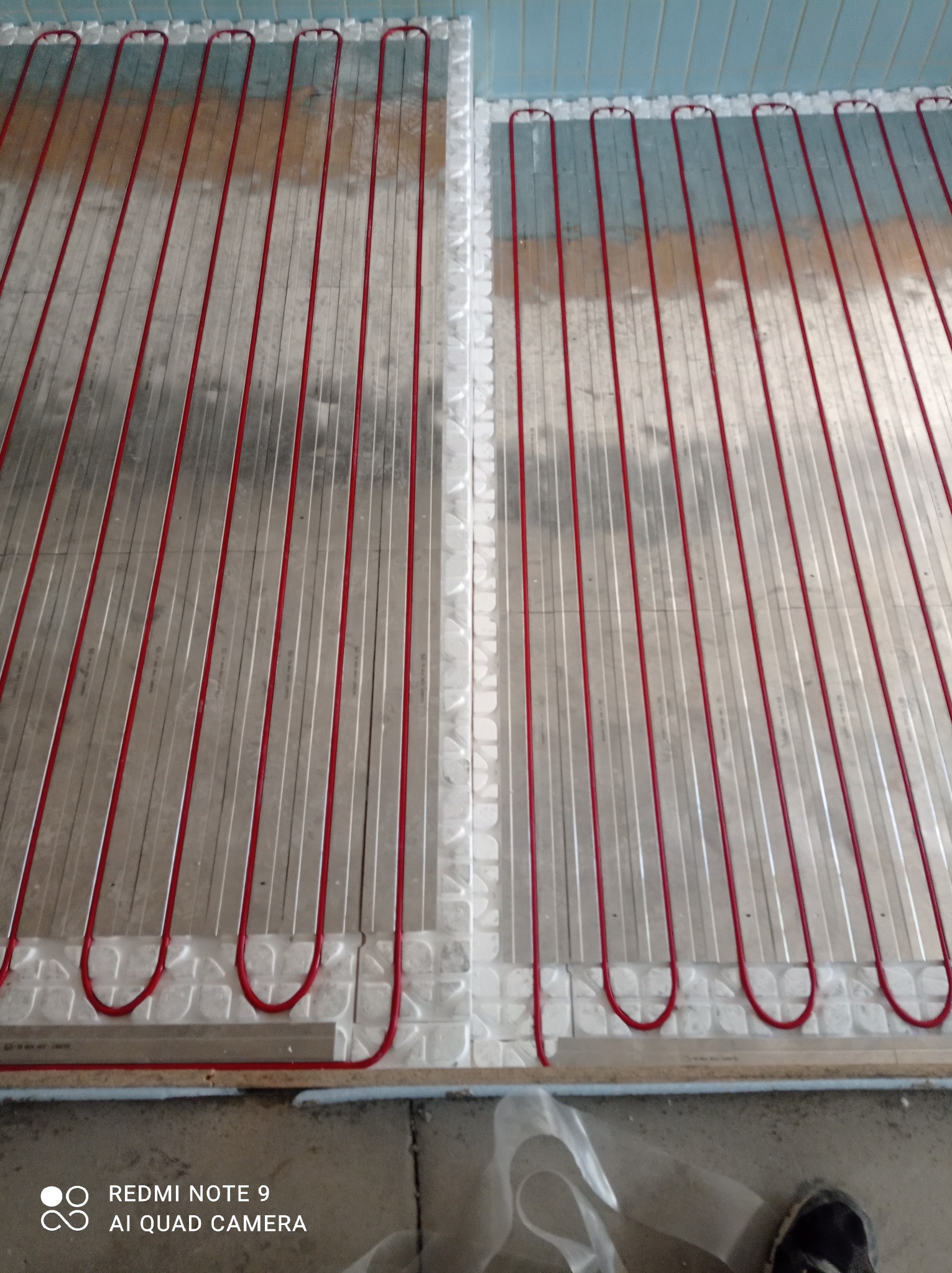 Czerwone rurki ogrzewania podłogowego ułożone na białej macie izolacyjnej, widoczne przed wylaniem wylewki, z fragmentem buta w prawym dolnym rogu.