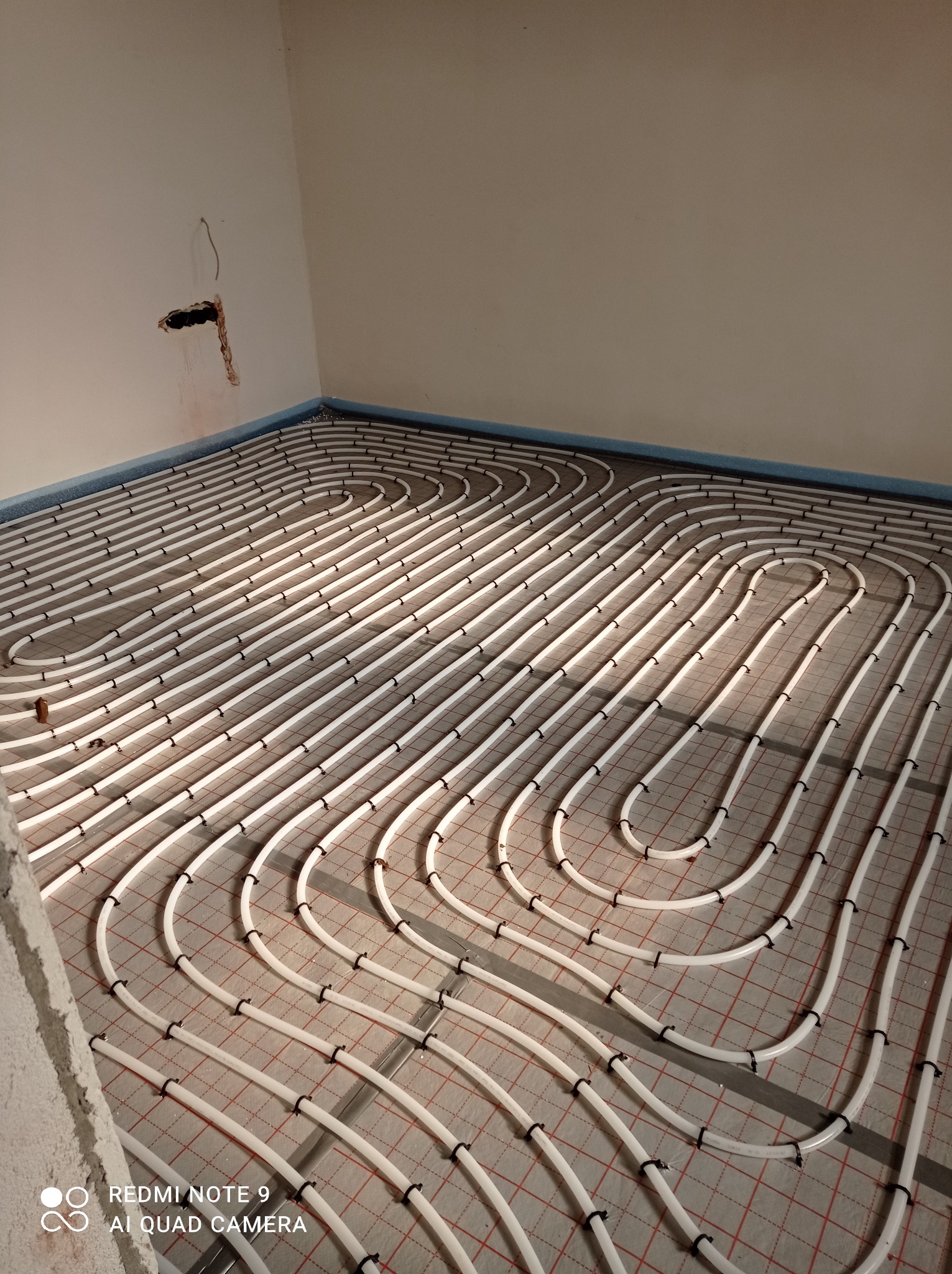 Białe rury ogrzewania podłogowego rozłożone na czerwonej siatce, widoczne mocowania, fragment ściany z otworem instalacyjnym w rogu pomieszczenia.