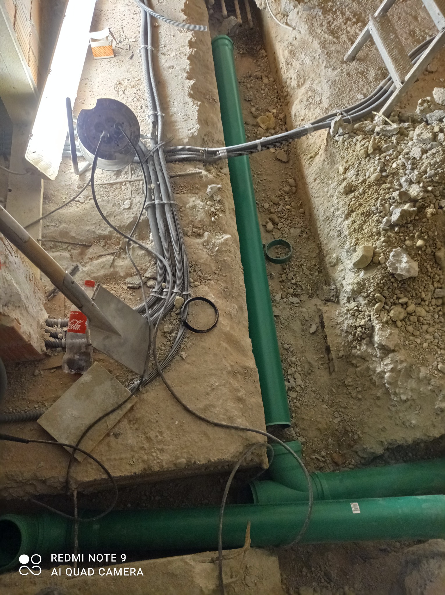 Wykop z ułożonymi rurami kanalizacyjnymi z tworzywa sztucznego, widoczne także kable elektryczne w peszlach, łopata i butelka Coca-Coli.