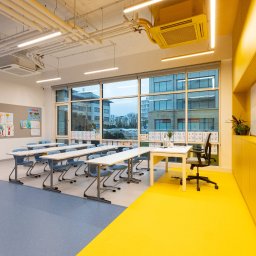 IWONA MAŁGORZATA CHMIELEWSKA PROFIT LINE - Wnętrze jasnej klasy szkolnej z dwukolorową podłogą: szara z drobnymi plamkami i żółta, biurka z niebieskimi krzesłami ustawione w rzędach, duże okna z widokiem na zabudowę miejską, tablica...