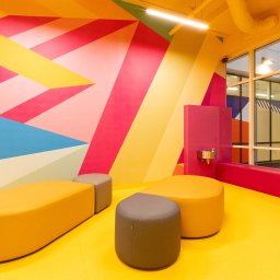IWONA MAŁGORZATA CHMIELEWSKA PROFIT LINE - Kolorowe wnętrze z geometrycznym muralem na ścianie, żółtą podłogą i pufami w odcieniach żółci i szarości, widoczny fragment okna z odbiciem muralu.