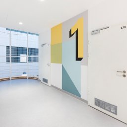 IWONA MAŁGORZATA CHMIELEWSKA PROFIT LINE - Korytarz w jasnych barwach z szarą, jednolitą posadzką, dekoracyjnym malowaniem ściany z motywem numeru jeden i białymi drzwiami z numerami pokoi.