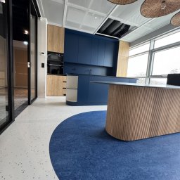 IWONA MAŁGORZATA CHMIELEWSKA PROFIT LINE - Nowoczesne wnętrze z kontrastującą podłogą: biała z drobnymi plamkami przechodzi w okrągłą, intensywnie niebieską strefę. W tle zabudowa kuchenna w odcieniach drewna i głębokiego granatu.