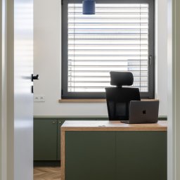 IWONA MAŁGORZATA CHMIELEWSKA PROFIT LINE - Nowoczesne biuro z drewnianą podłogą w jodełkę, zielonymi szafkami i biurkiem z laptopem, widok zza otwartych drzwi.