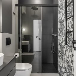 IWONA MAŁGORZATA CHMIELEWSKA PROFIT LINE - Nowoczesna łazienka z czarno-białą tapetą w roślinne wzory, kabina prysznicowa ze szklanymi drzwiami przesuwnymi, umywalka nablatowa i czarne płytki ścienne.