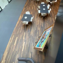 IWONA MAŁGORZATA CHMIELEWSKA PROFIT LINE - Widok z góry na biurową przestrzeń z podłogą z ciemnego drewna, stołami i krzesłami, oraz obrazem opartym o ścianę.