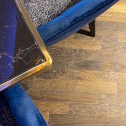 IWONA MAŁGORZATA CHMIELEWSKA PROFIT LINE - Fragment wnętrza: brązowa podłoga z paneli, fragment niebieskiego fotela z weluru i stolika z marmurowym blatem i złotą ramą.
