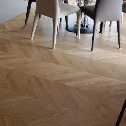 Parkiet ułożony we wz&oacute;r jodełki francuskiej, widoczne krzesła i fragment stołu w tle, w rogu kratka wentylacyjna.