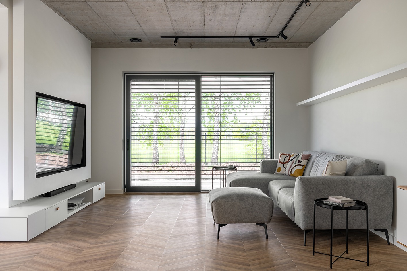 Salon z parkietem ułożonym we wzór jodełki, szarą sofą i widokiem na zieleń za oknem z żaluzjami.