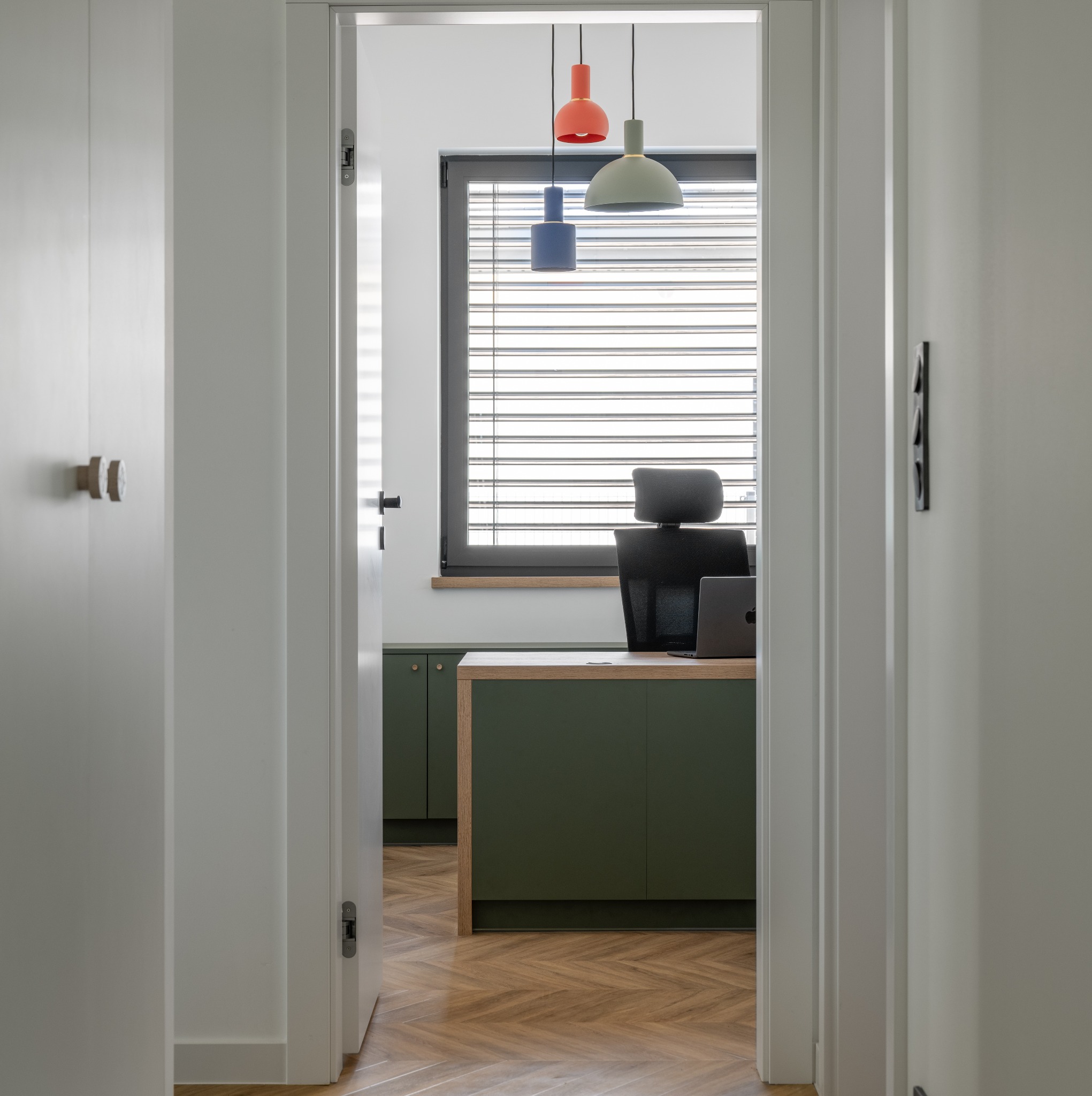 Wnętrze biura domowego z podłogą ułożoną w jodełkę, widok przez otwarte białe drzwi, z trzema wiszącymi lampami w kolorach pomarańczowym, zielonym i niebieskim nad biurkiem z laptopem i krzesłem...