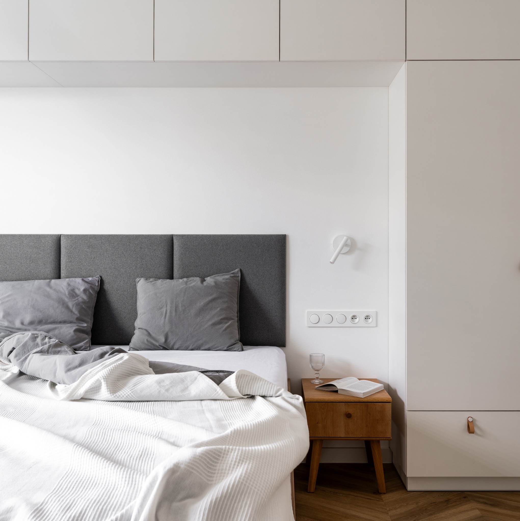 Minimalistyczna sypialnia z parkietem w jodełkę, szarą tapicerowaną ścianą za łóżkiem, białą pościelą i szafką nocną z książką i szklanką.