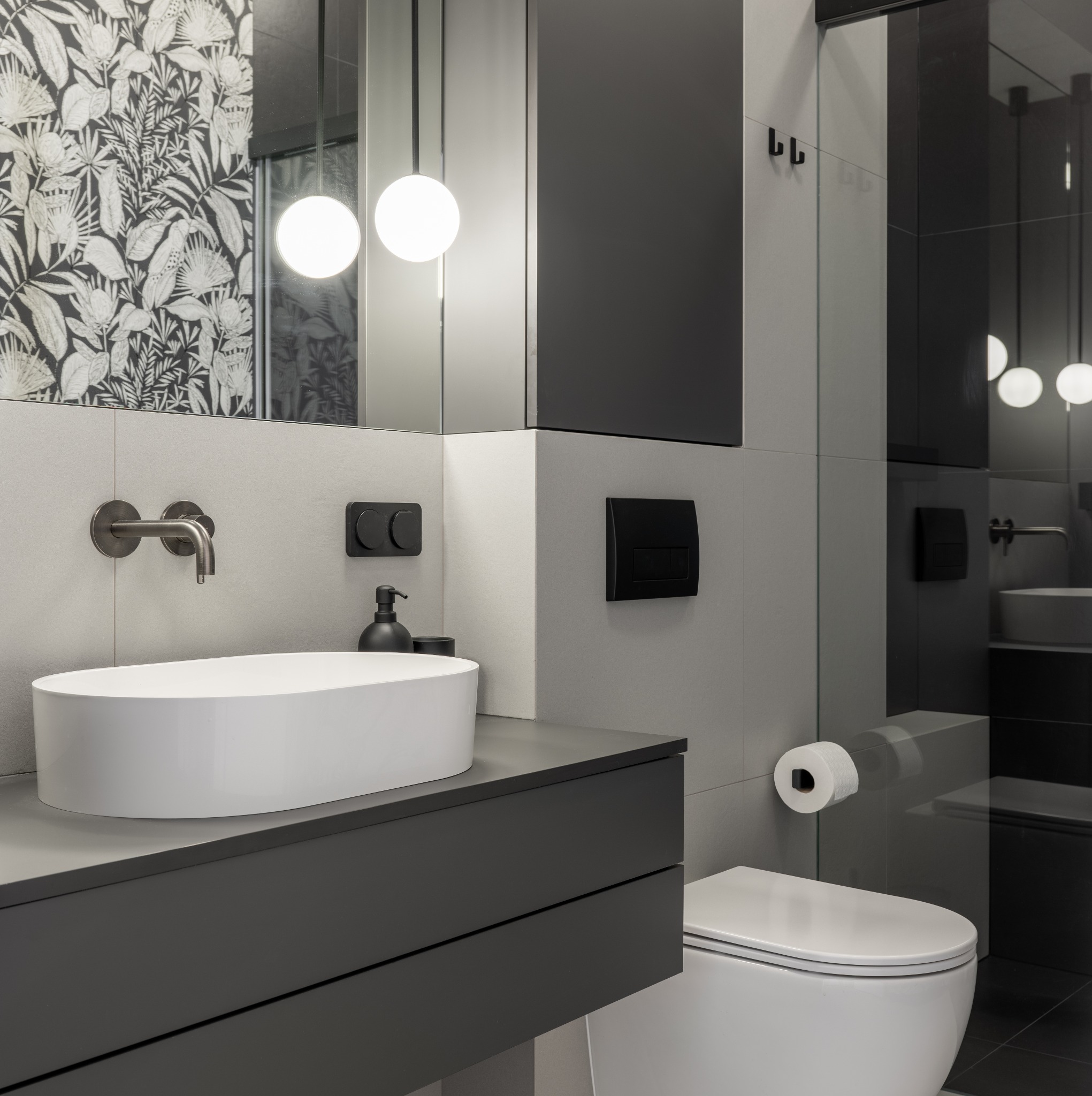 Nowoczesna łazienka w szarościach z białą umywalką nablatową i toaletą, wzorzysta tapeta w tle, minimalistyczna armatura i oświetlenie kuliste.