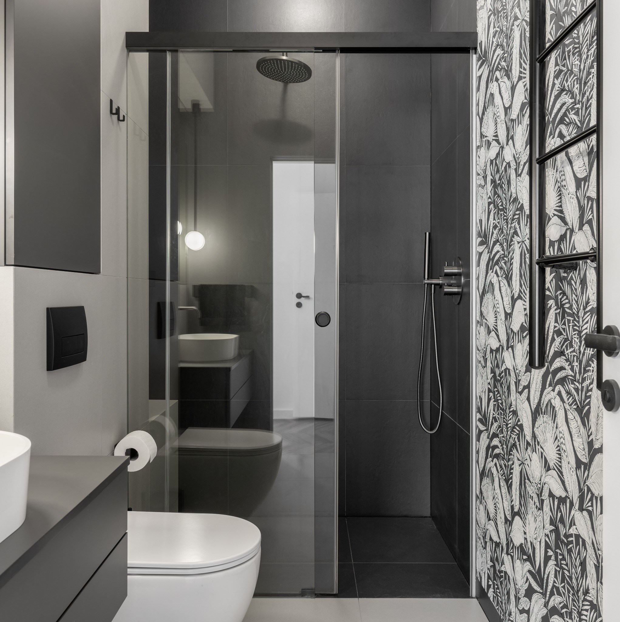 Nowoczesna łazienka z czarno-białą tapetą w roślinne wzory, kabina prysznicowa ze szklanymi drzwiami przesuwnymi, umywalka nablatowa i czarne płytki ścienne.