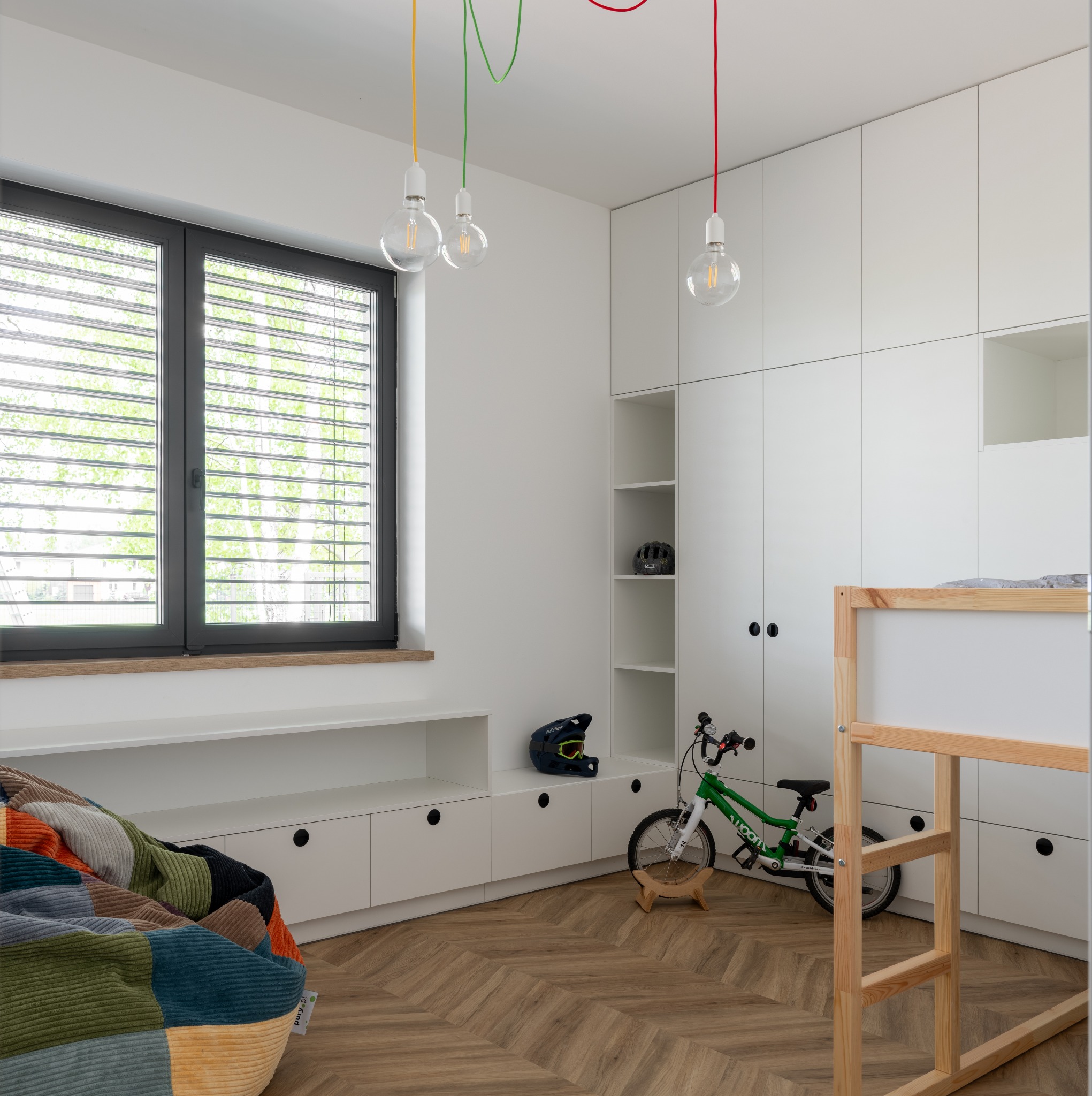 Pokój dziecięcy z drewnianą podłogą ułożoną we wzór jodełki, białymi szafkami i łóżkiem piętrowym, oświetlony wiszącymi lampami z kolorowymi kablami.