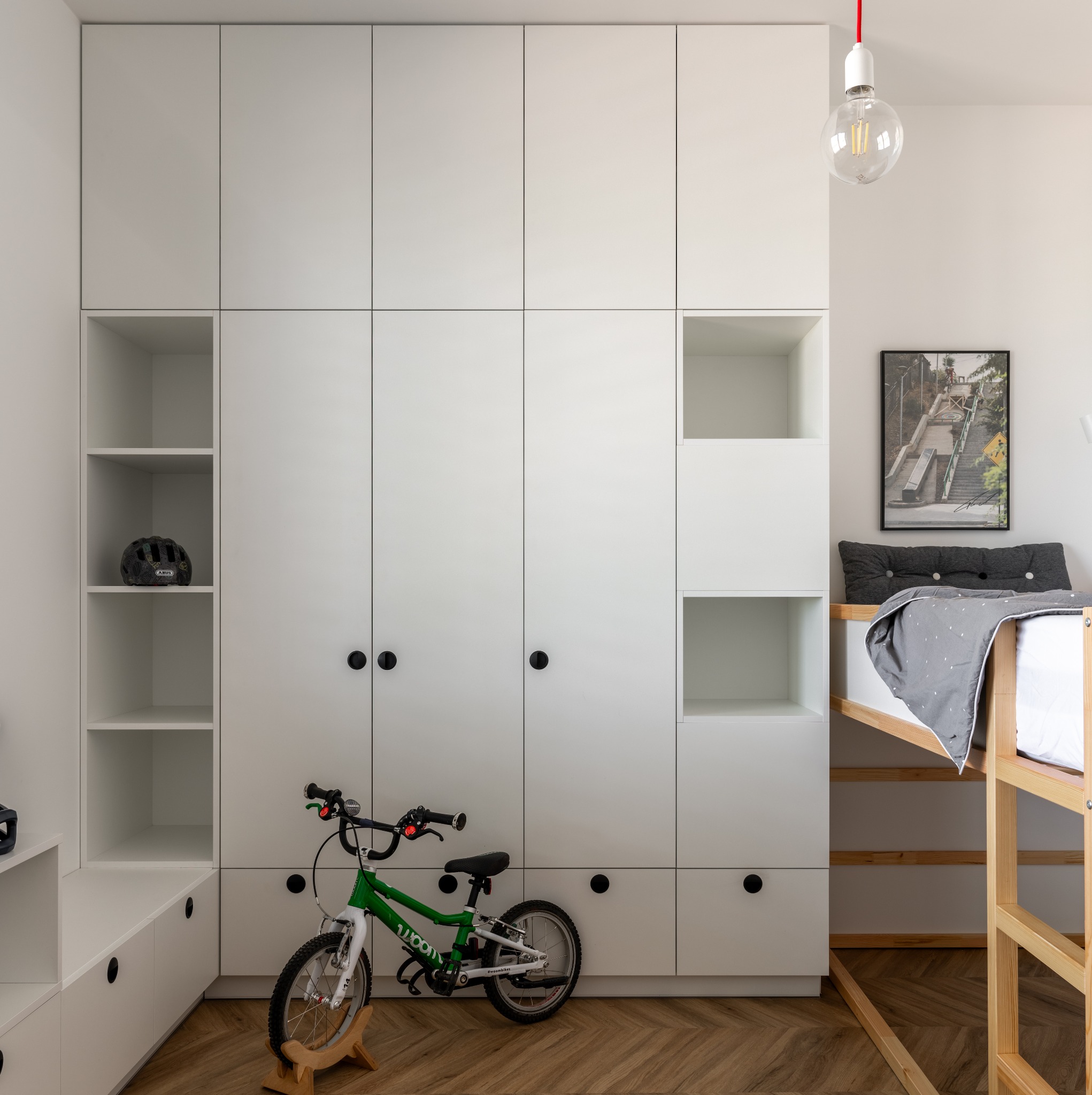 Nowoczesny pokój dziecięcy z białymi szafami w zabudowie, drewnianym łóżkiem piętrowym i parkietem w jodełkę, zielony rower dziecięcy oparty o szafę, na półce czarny kask.