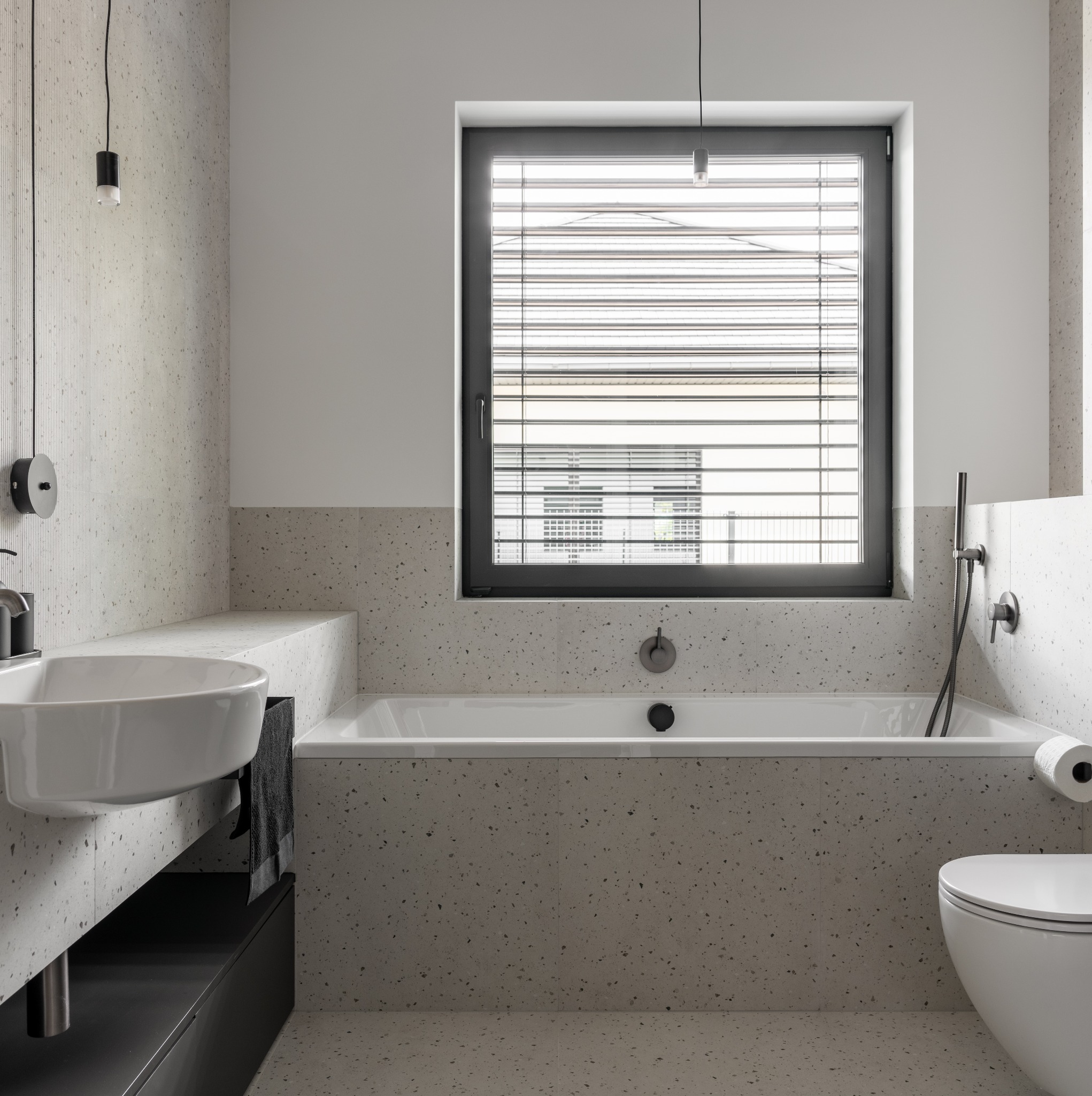 Nowoczesna łazienka z wanną, umywalką i toaletą, wykończona płytkami imitującymi lastryko, z oknem z żaluzjami i minimalistycznym oświetleniem.