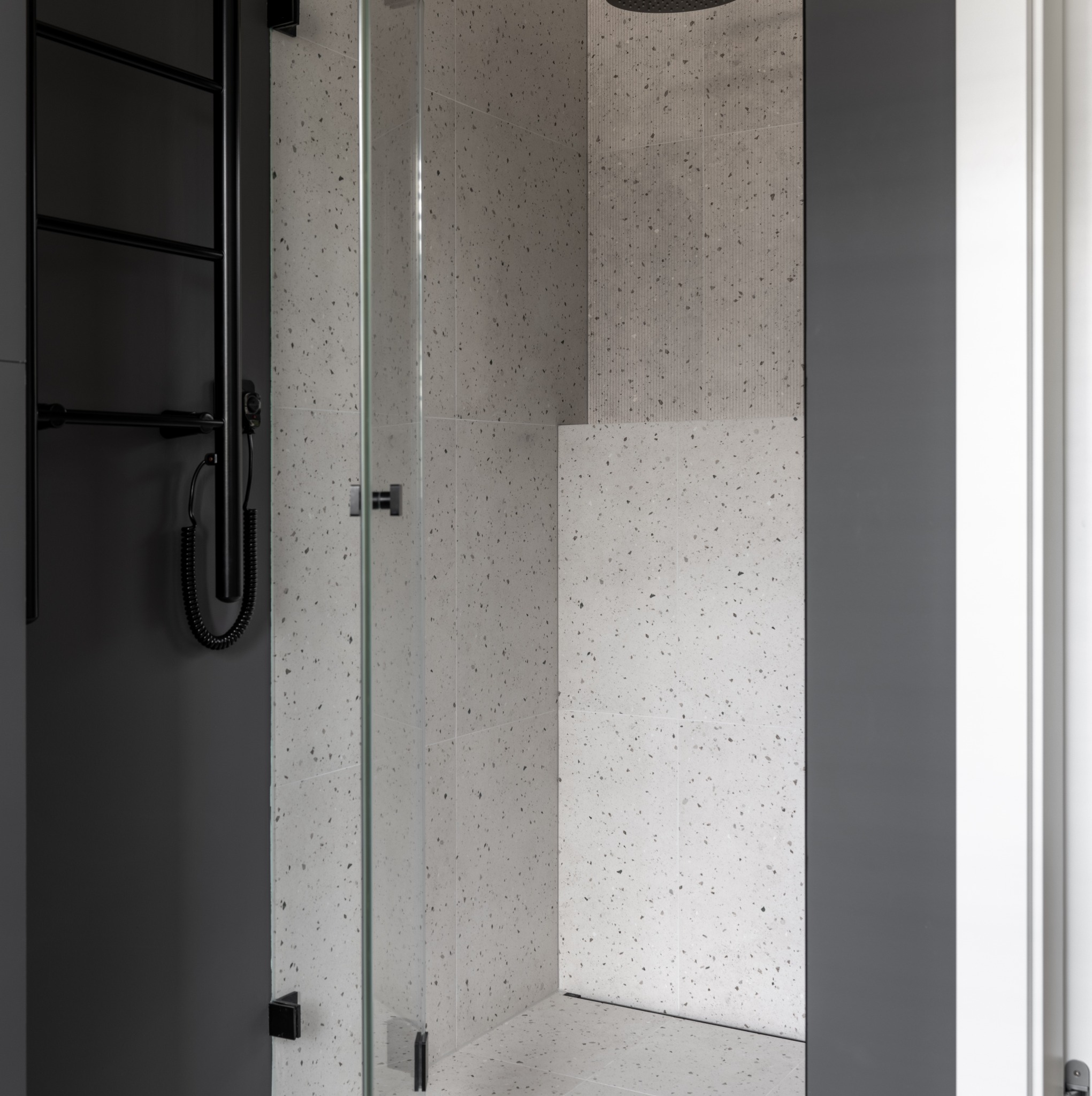 Nowoczesna kabina prysznicowa z szklanymi drzwiami, wykończona płytkami w odcieniach szarości z wzorem lastryko, czarny grzejnik drabinkowy na ścianie.