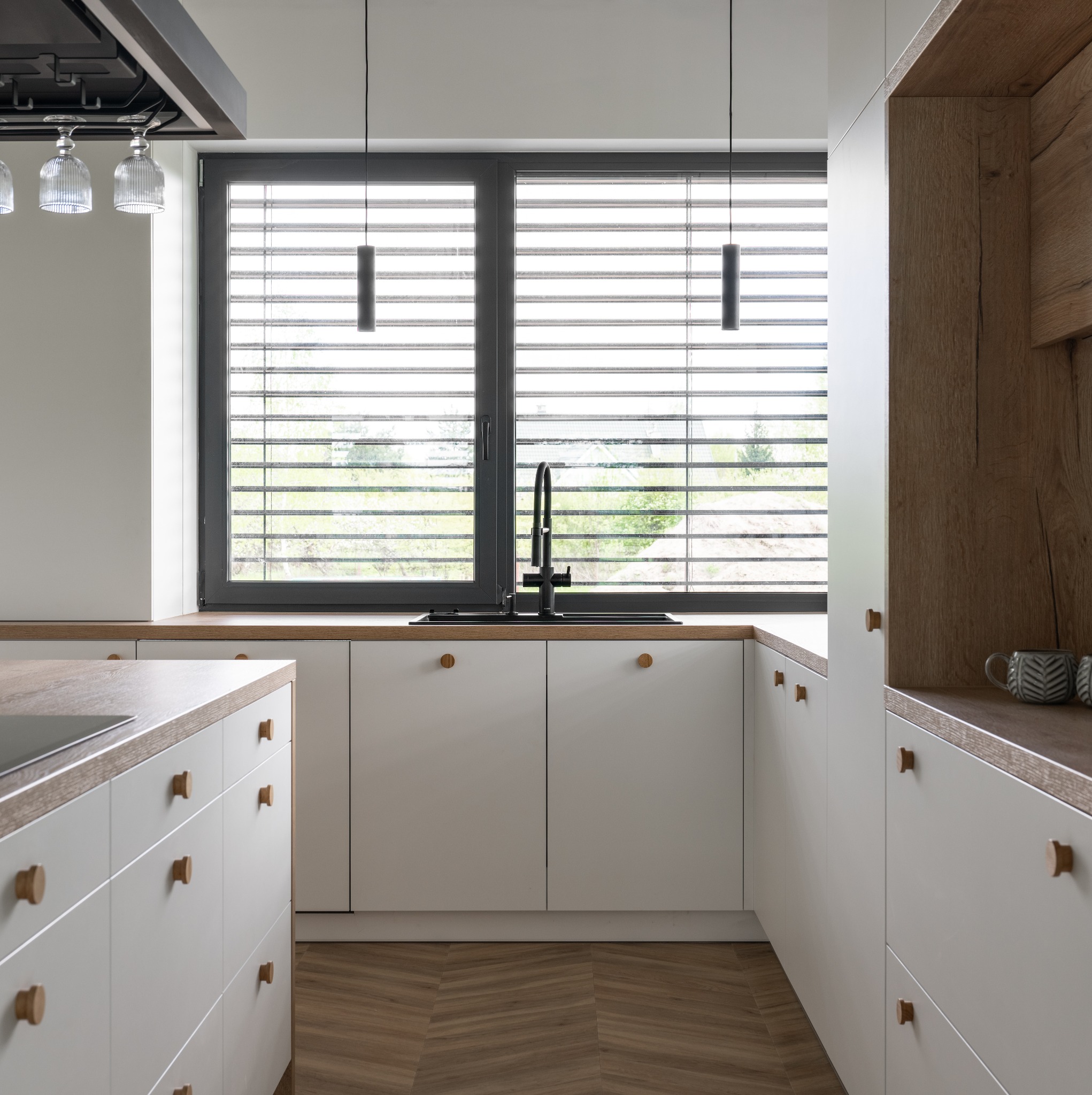 Nowoczesna kuchnia z białymi szafkami, drewnianymi blatami i podłogą w jodełkę, widok na okno z żaluzjami.