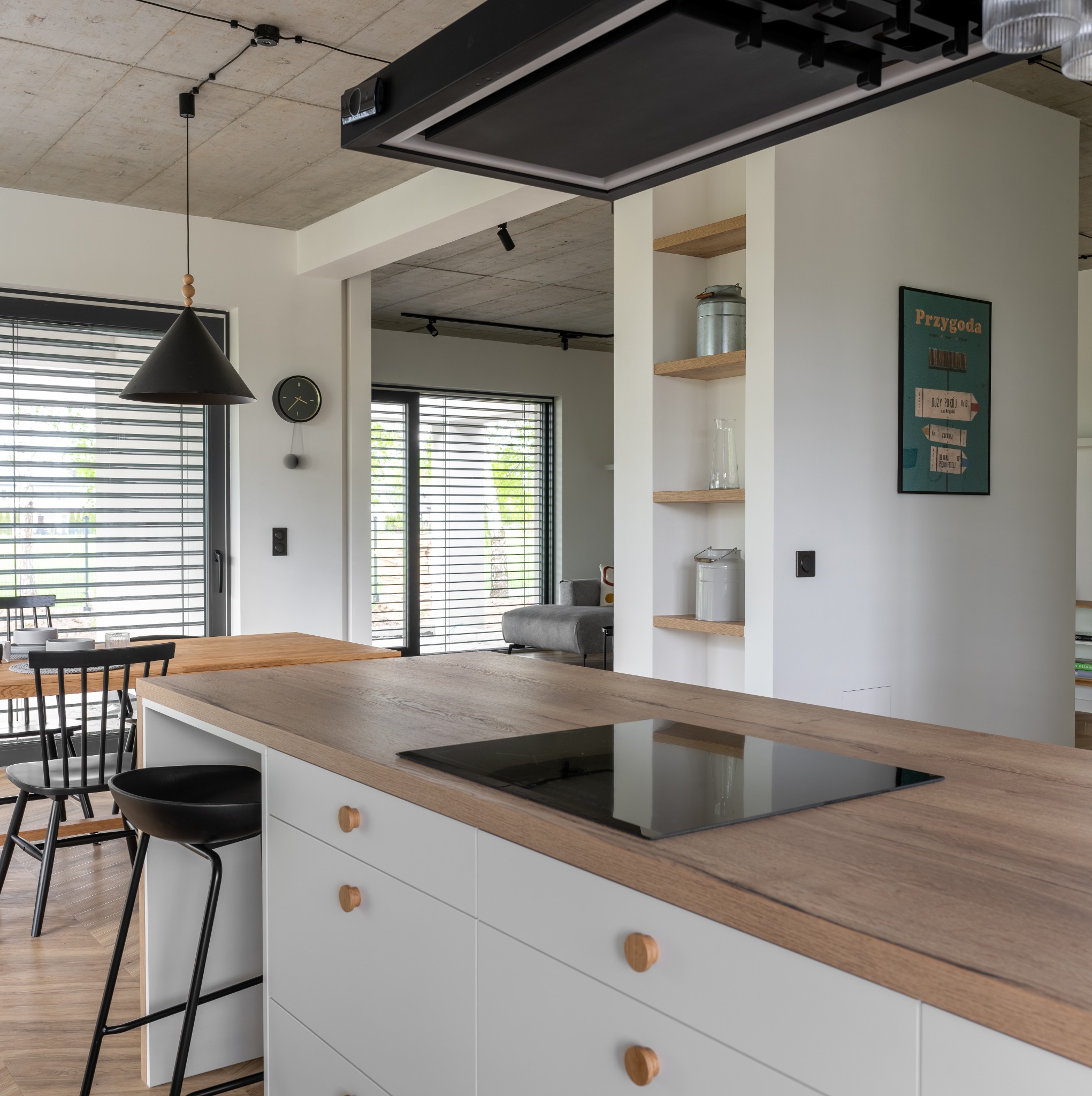 Nowoczesna kuchnia z drewnianym blatem, czarną płytą indukcyjną, białymi szafkami z okrągłymi uchwytami i czarnym stołkiem barowym na tle parkietu ułożonego w jodełkę.