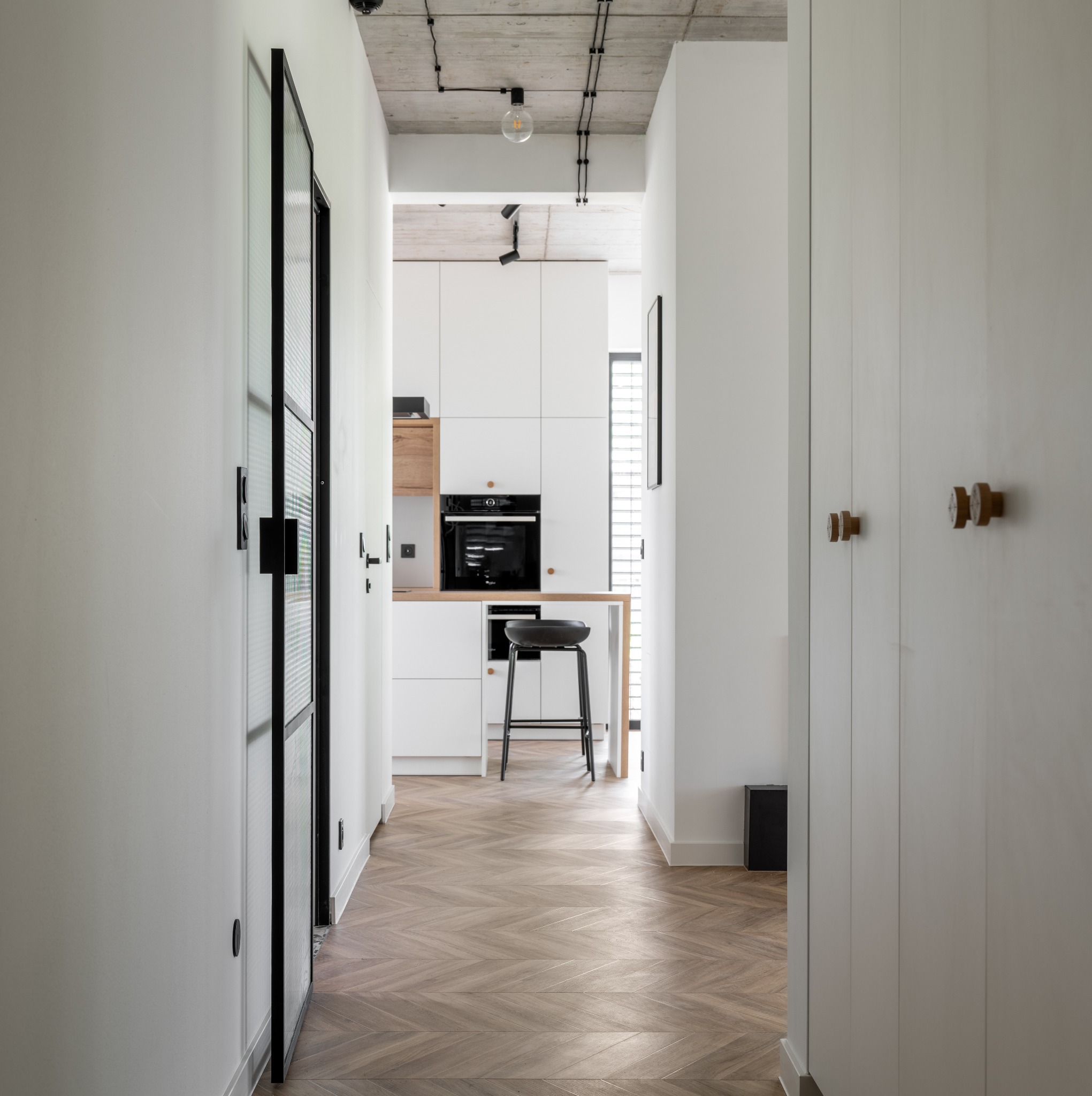 Korytarz z parkietem ułożonym we wzór jodełki francuskiej, widok na kuchnię z białymi szafkami i czarnym stołkiem barowym.