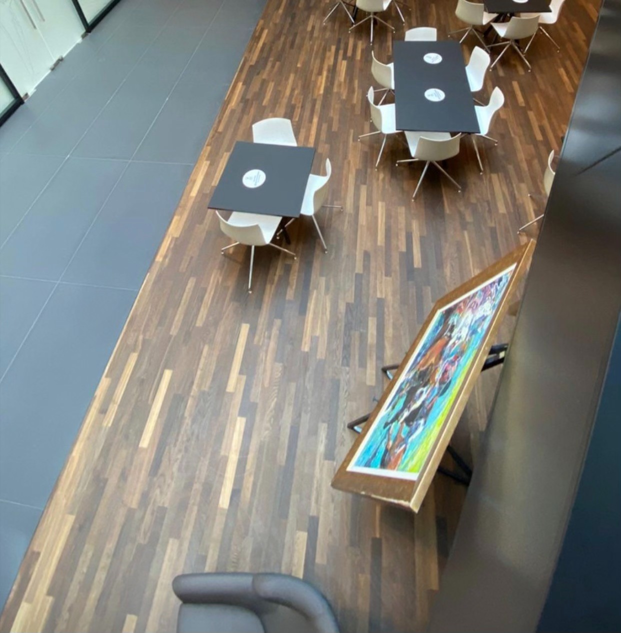 Widok z góry na biurową przestrzeń z podłogą z ciemnego drewna, stołami i krzesłami, oraz obrazem opartym o ścianę.