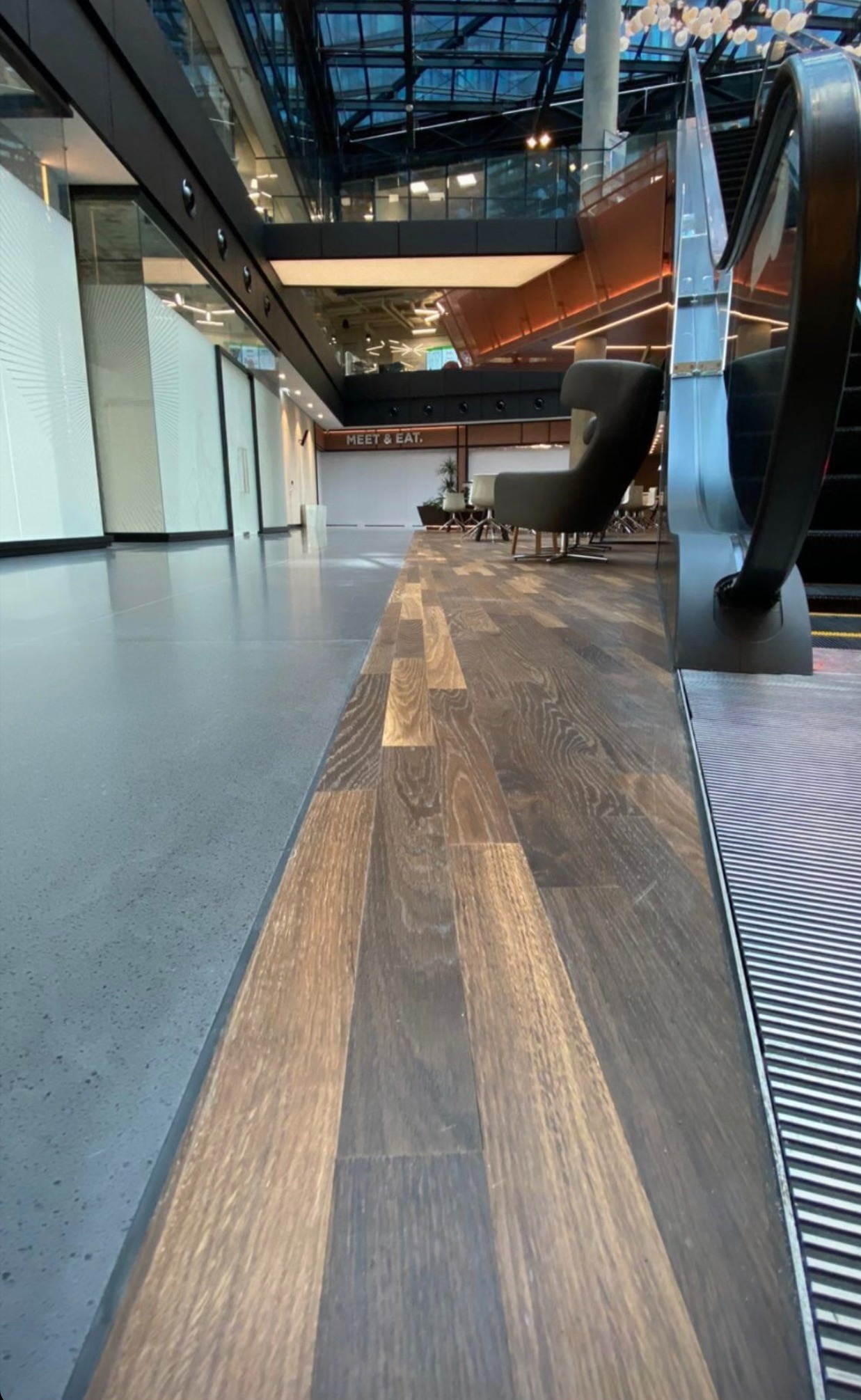 Parkiet z ciemnego drewna ułożony wzdłuż ruchomych schodów w nowoczesnym wnętrzu budynku z przeszklonym sufitem i jasnymi ścianami.