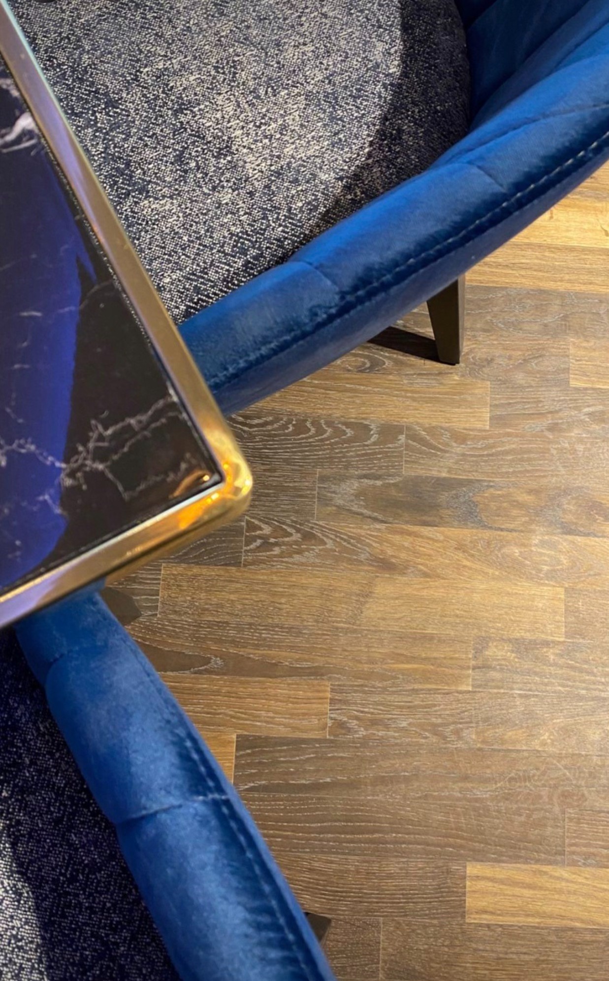 Fragment wnętrza: brązowa podłoga z paneli, fragment niebieskiego fotela z weluru i stolika z marmurowym blatem i złotą ramą.
