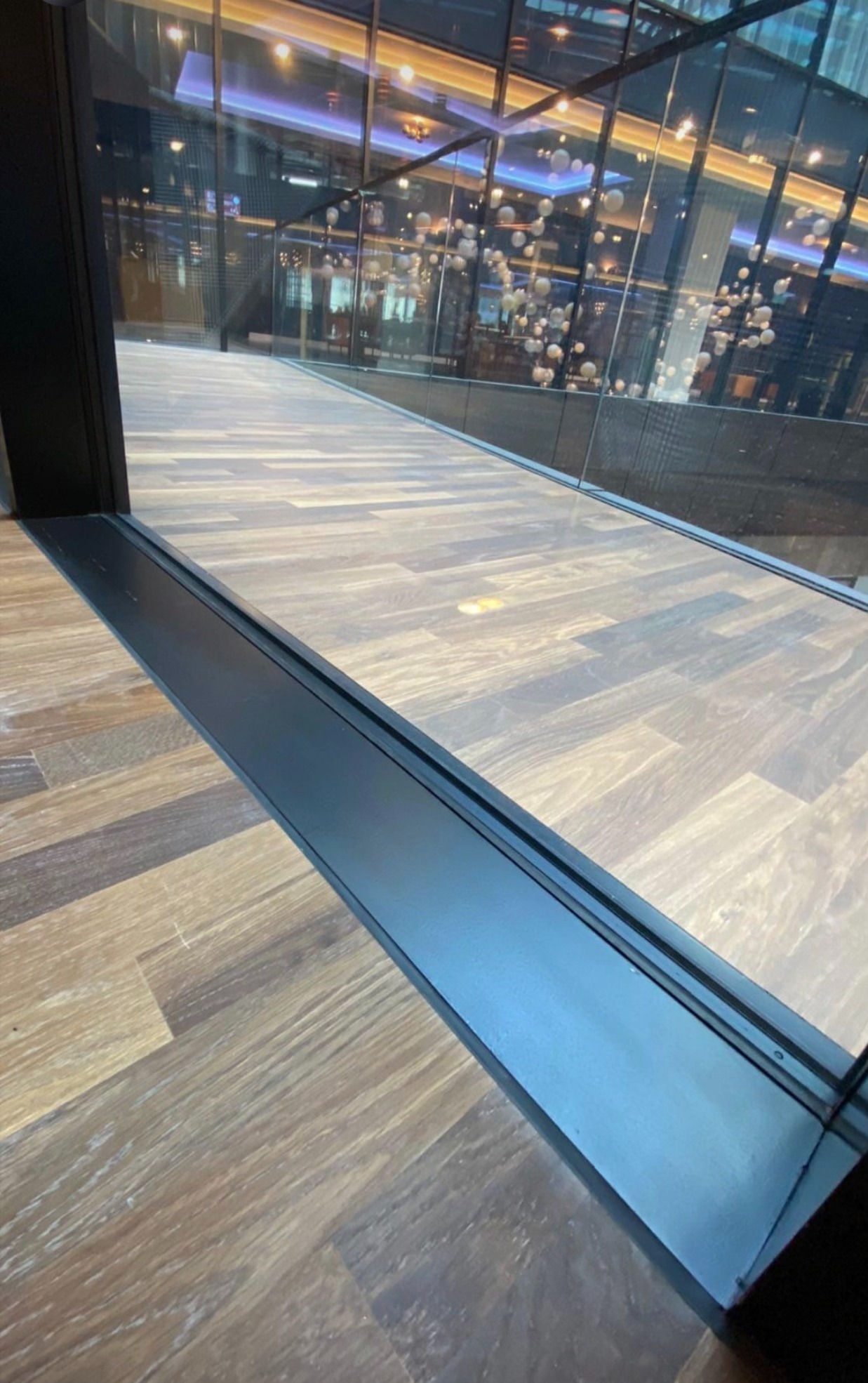 Wnętrze nowoczesnego biurowca z podłogą imitującą drewno, widoczne przez szklaną ścianę z czarną ramą.