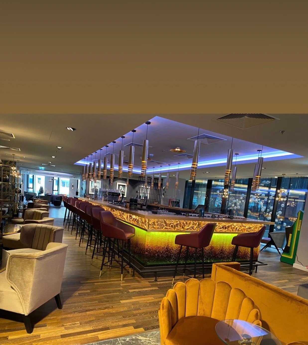 Wnętrze baru z drewnianą podłogą w jodełkę, podświetlonym blatem i krzesłami barowymi w kolorze burgundu, widok na salon z fotelami w odcieniach złota.