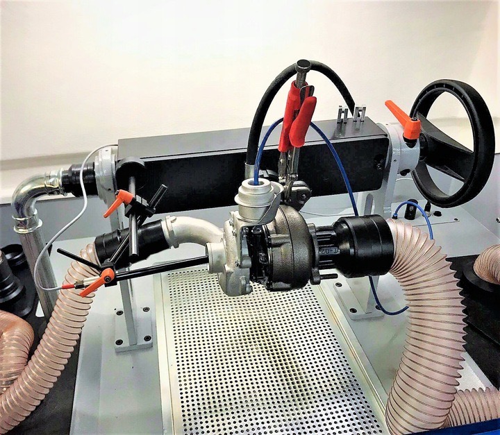 Turbosprężarka zamontowana na stanowisku testowym z podłączonymi przewodami i zaciskami, widoczna perforowana podstawa i elementy regulacyjne.