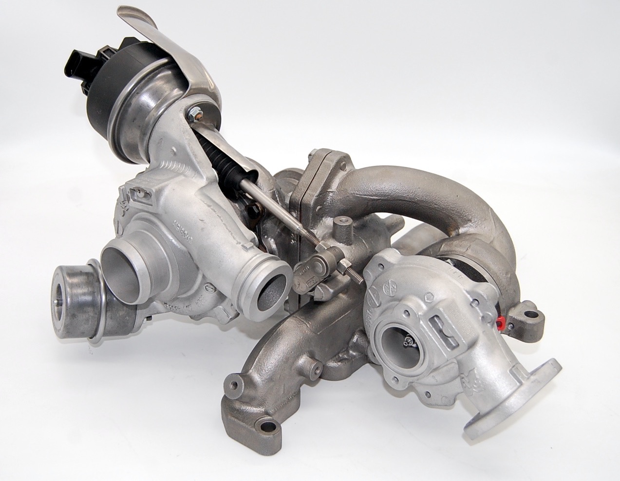 Turbosprężarka samochodowa, widoczne elementy metalowe i plastikowe, ustawiona na białym tle, oświetlenie studyjne.