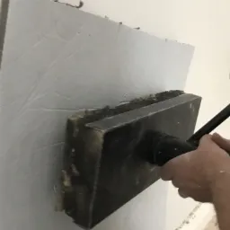 Aplikacja szarej masy szpachlowej na ścianę za pomocą dużej pacy z uchwytem, widoczne nierówności i ślady po narzędziu.