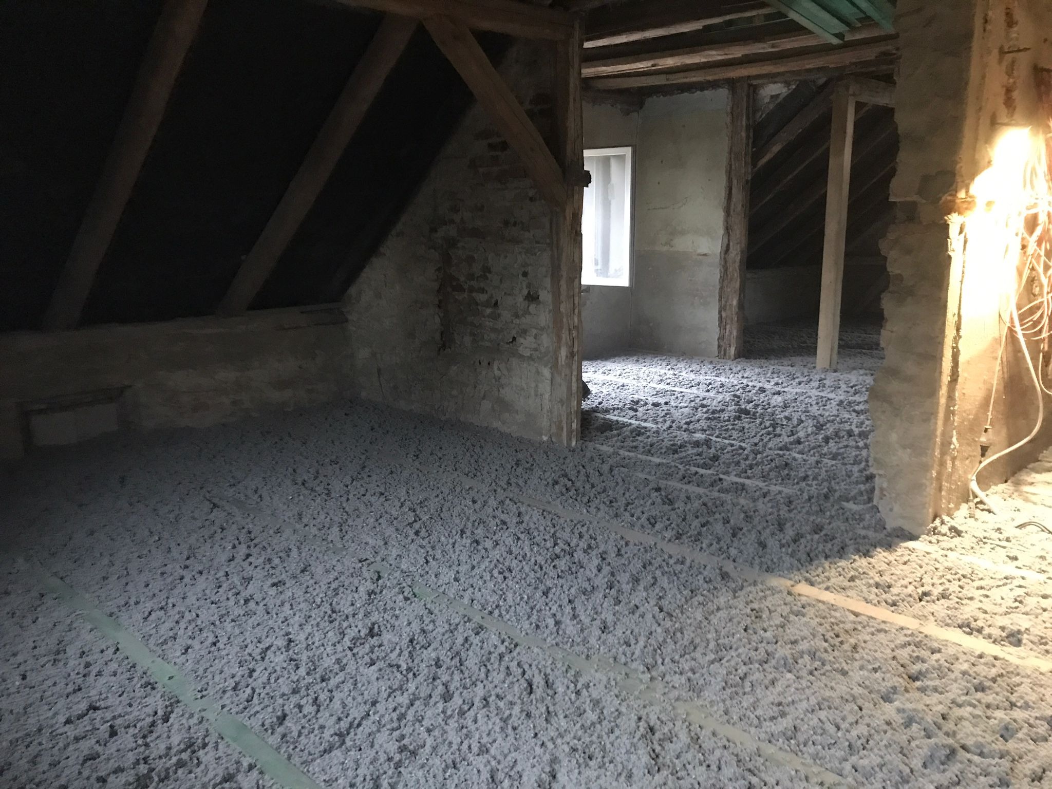 Poddasze z widoczną warstwą izolacji wdmuchiwanej z celulozy, rozłożoną równomiernie na podłodze między legarami, surowe ściany i drewniane elementy konstrukcyjne.