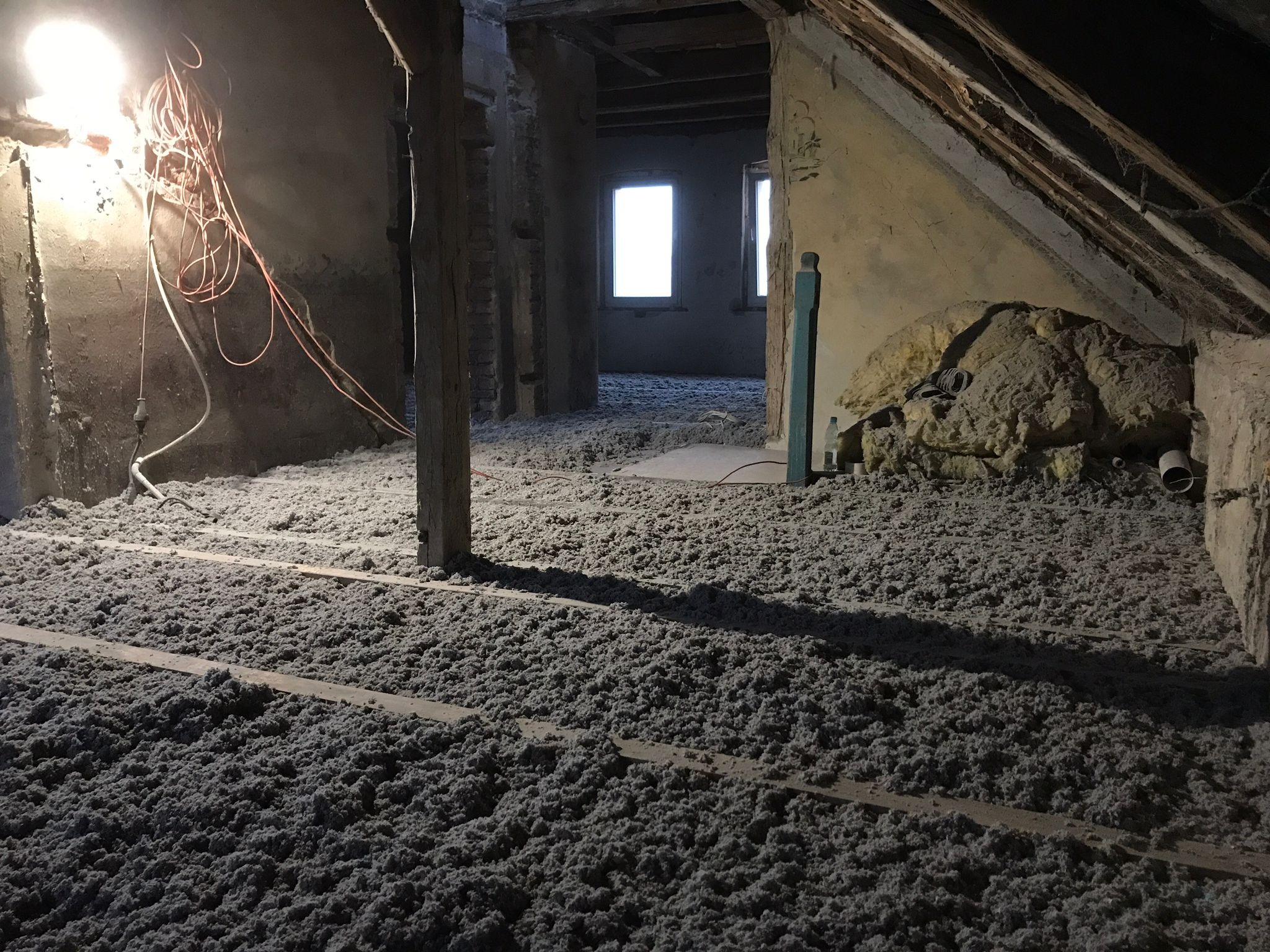 Wnętrze poddasza z widoczną warstwą szarej, wdmuchiwanej termoizolacji z celulozy, ułożonej między drewnianymi legarami; widoczne stare ściany i okna.