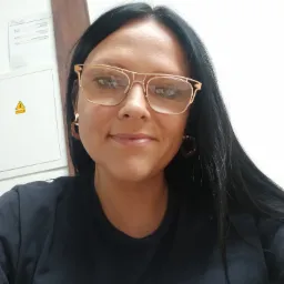 Kobieta w okularach z metalową oprawką i ciemnych włosach, uśmiechnięta, z widocznym symbolem planety na koszulce, stojąca na tle białej ściany z szafką elektryczną i znakiem ostrzegawczym.