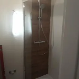 Nowa kabina prysznicowa z panelem imitującym drewno, widoczne przyłącza wodne na ścianie i poziomica z boku.