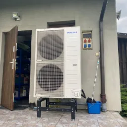 Biała pompa ciepła Samsung zamontowana na czarnym stelażu przed szarą ścianą budynku z otwartymi drzwiami garażowymi i niebieskim wiadrem z mopem obok rury spustowej.