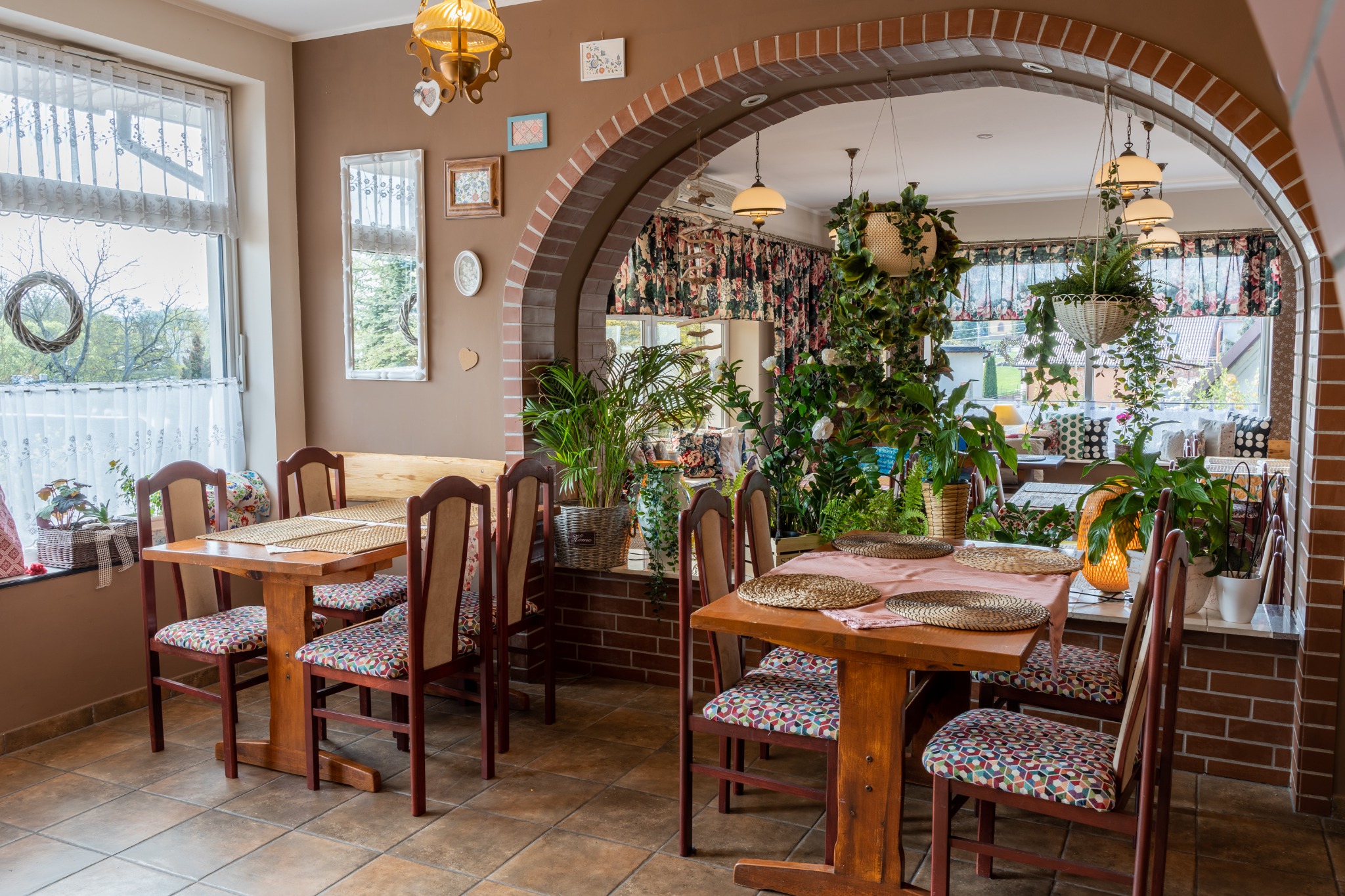 Urokliwe wnętrze restauracji z drewnianymi stołami nakrytymi plecionymi podkładkami, krzesła z geometrycznym wzorem, ceglany łuk prowadzący do drugiej części sali, dekoracje roślinne i rustykalne...