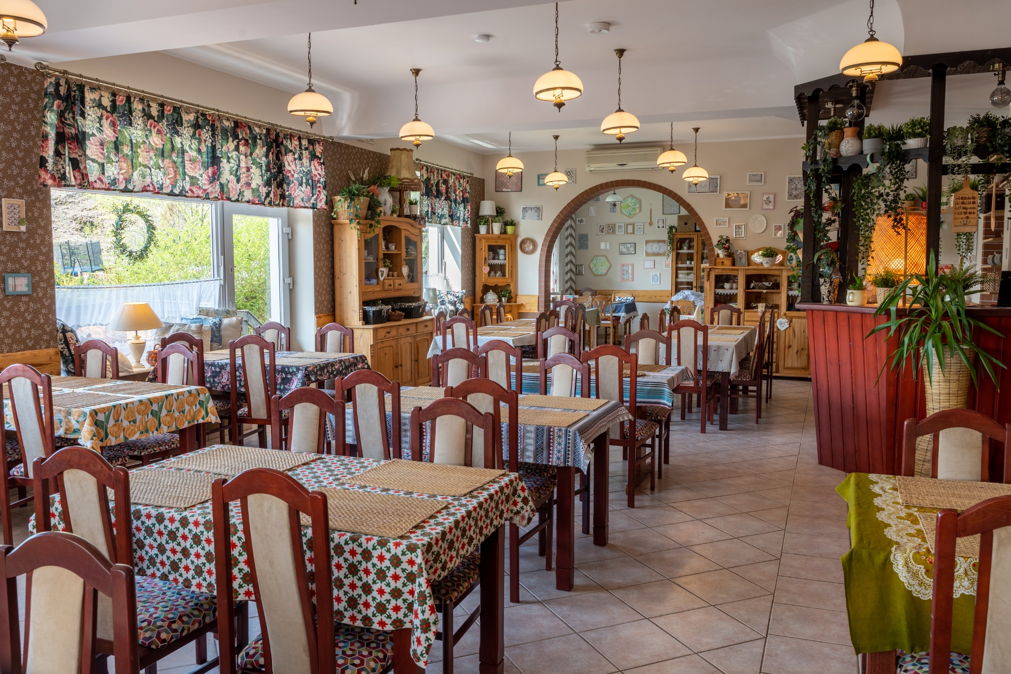 Sala restauracyjna z nakrytymi stołami, dekoracjami roślinnymi i klasycznymi lampami wiszącymi, gotowa na przyjęcie gości.