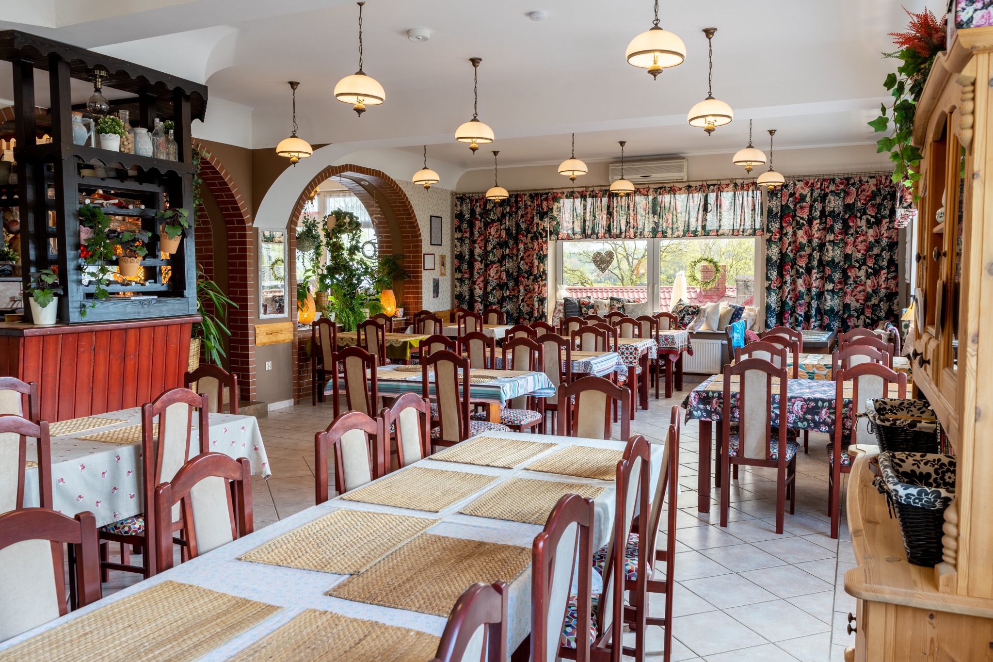 Jasne wnętrze restauracji z nakrytymi stołami, drewnianymi krzesłami i dekoracyjnymi elementami, takimi jak kwiatowe zasłony i wiszące lampy.