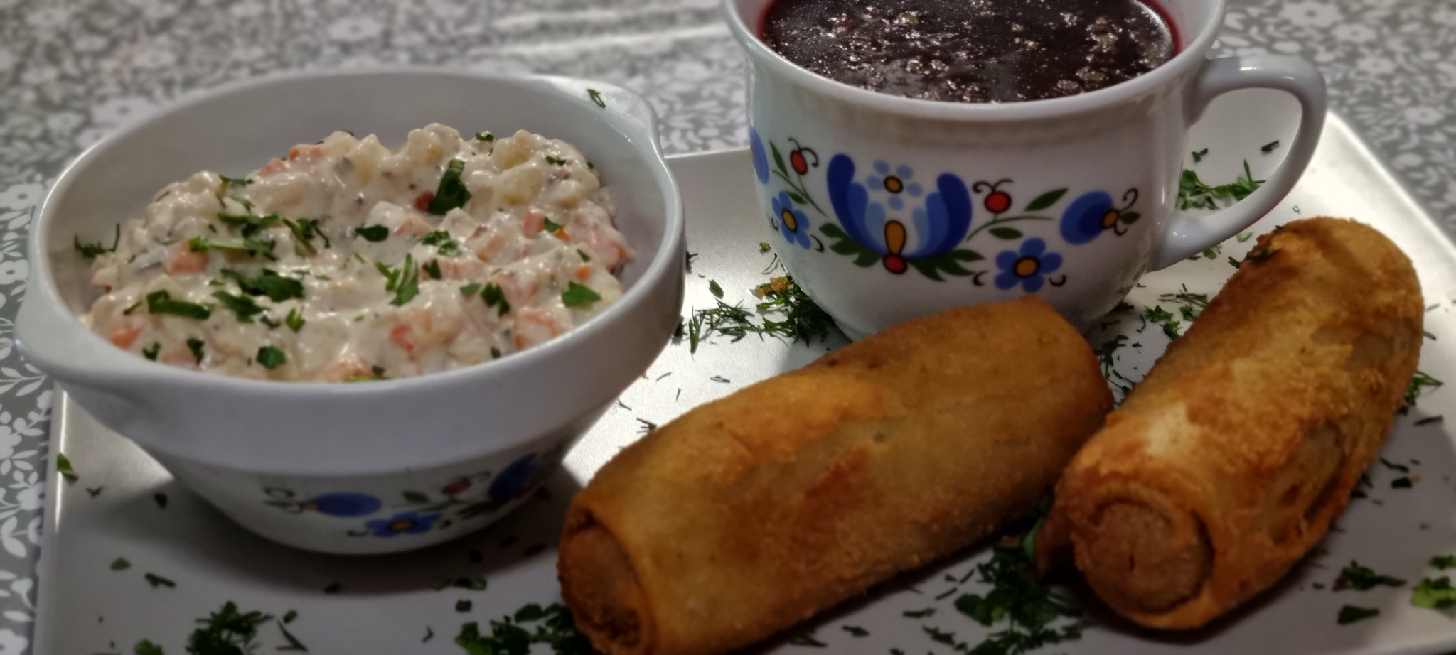 Tradycyjne polskie dania: sałatka jarzynowa z majonezem w białej miseczce, barszcz czerwony w kubku z folkowym wzorem oraz dwa krokiety na białym talerzu, posypane koperkiem.