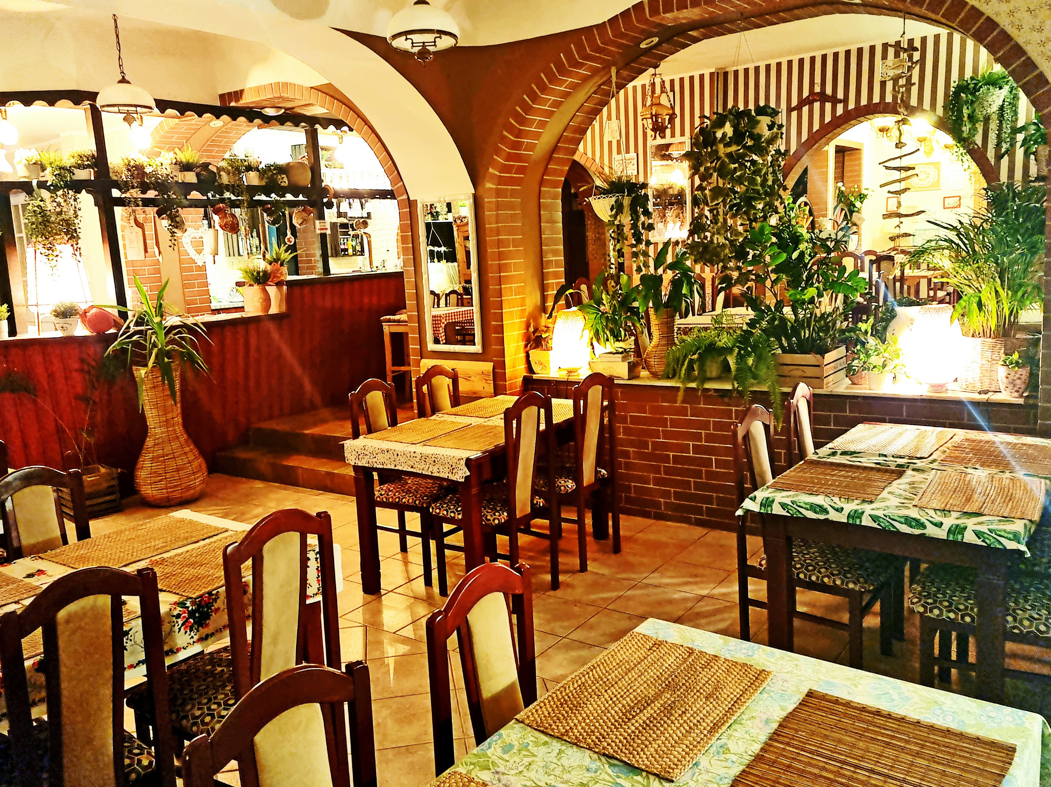 Ustawienie stołów w restauracji z ceglanymi łukami, roślinami doniczkowymi i ozdobnymi lampami, tworzące przytulną atmosferę.