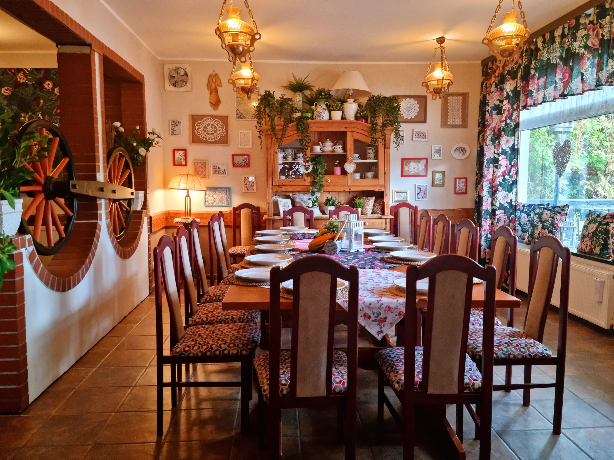 Uroczysta aranżacja długiego stołu z nakryciami, kwiatowym obrusem i dekoracjami, w rustykalnym wnętrzu z drewnianą komodą, oświetlonym wiszącymi lampami.