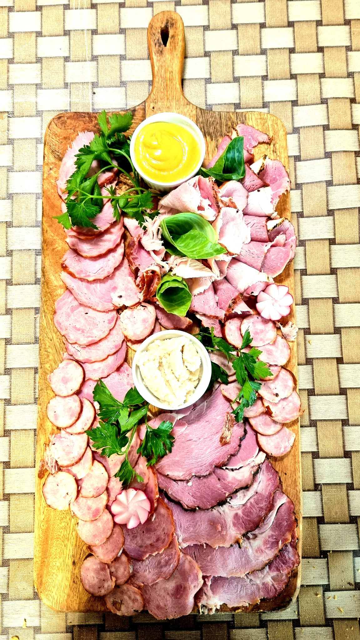 Deska wędlin i kiełbas z dipami i dekoracją z zieleniny, przygotowana przez firmę z Chmielna, na tle plecionej maty.