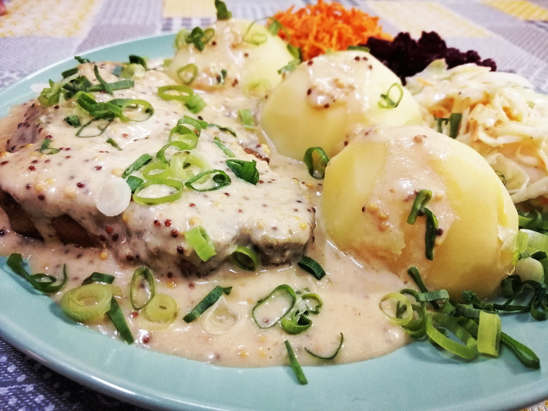 Gotowe danie na turkusowym talerzu: mięso w sosie musztardowym, ziemniaki, surówka z marchwi i kapusty, posypane zielonym szczypiorkiem.