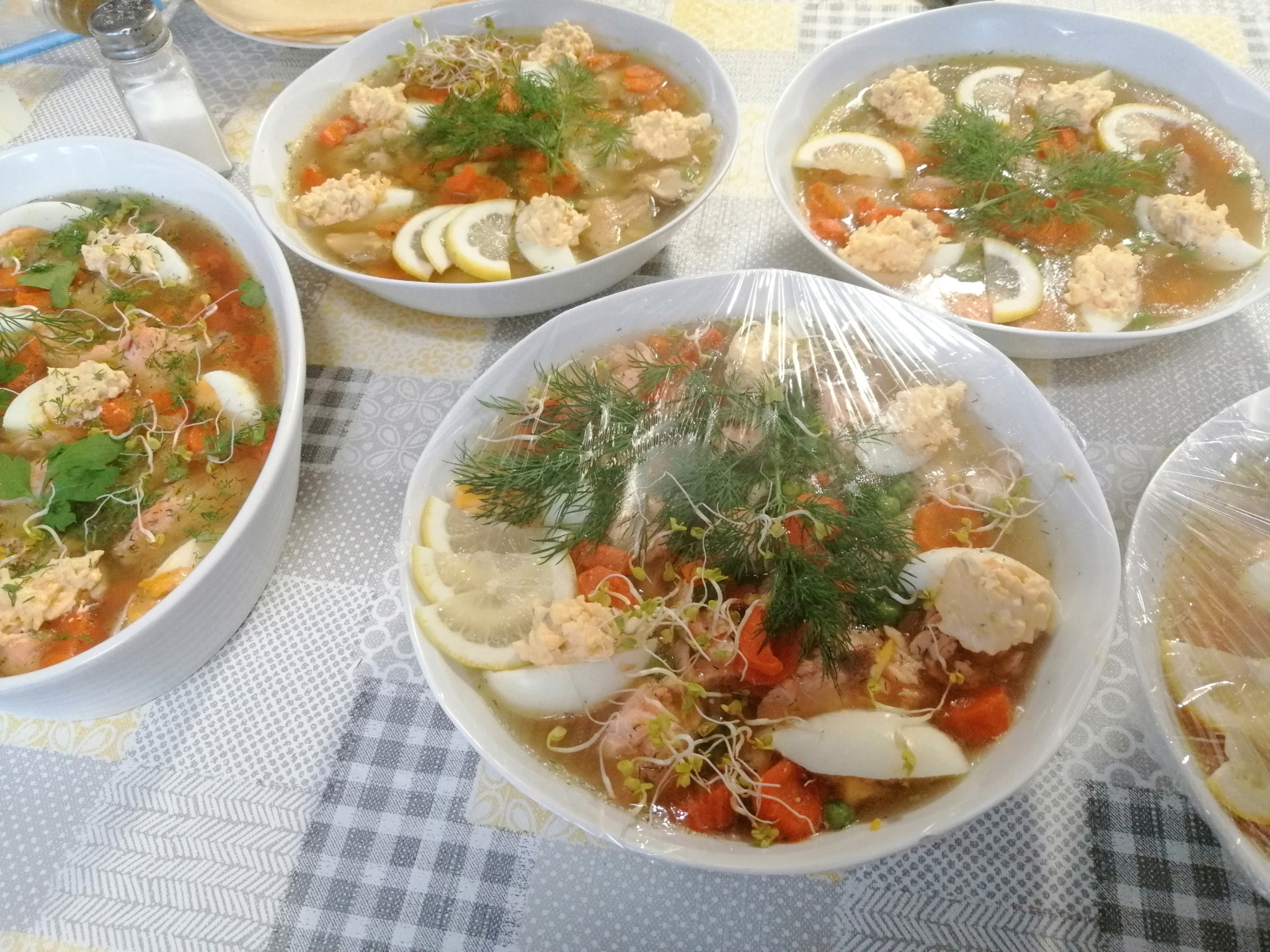 Cztery białe miski z galaretą rybną, jajkiem, marchewką, cytryną, koperkiem i kiełkami na wzorzystym obrusie, jedna miska owinięta folią spożywczą, w tle solniczka.