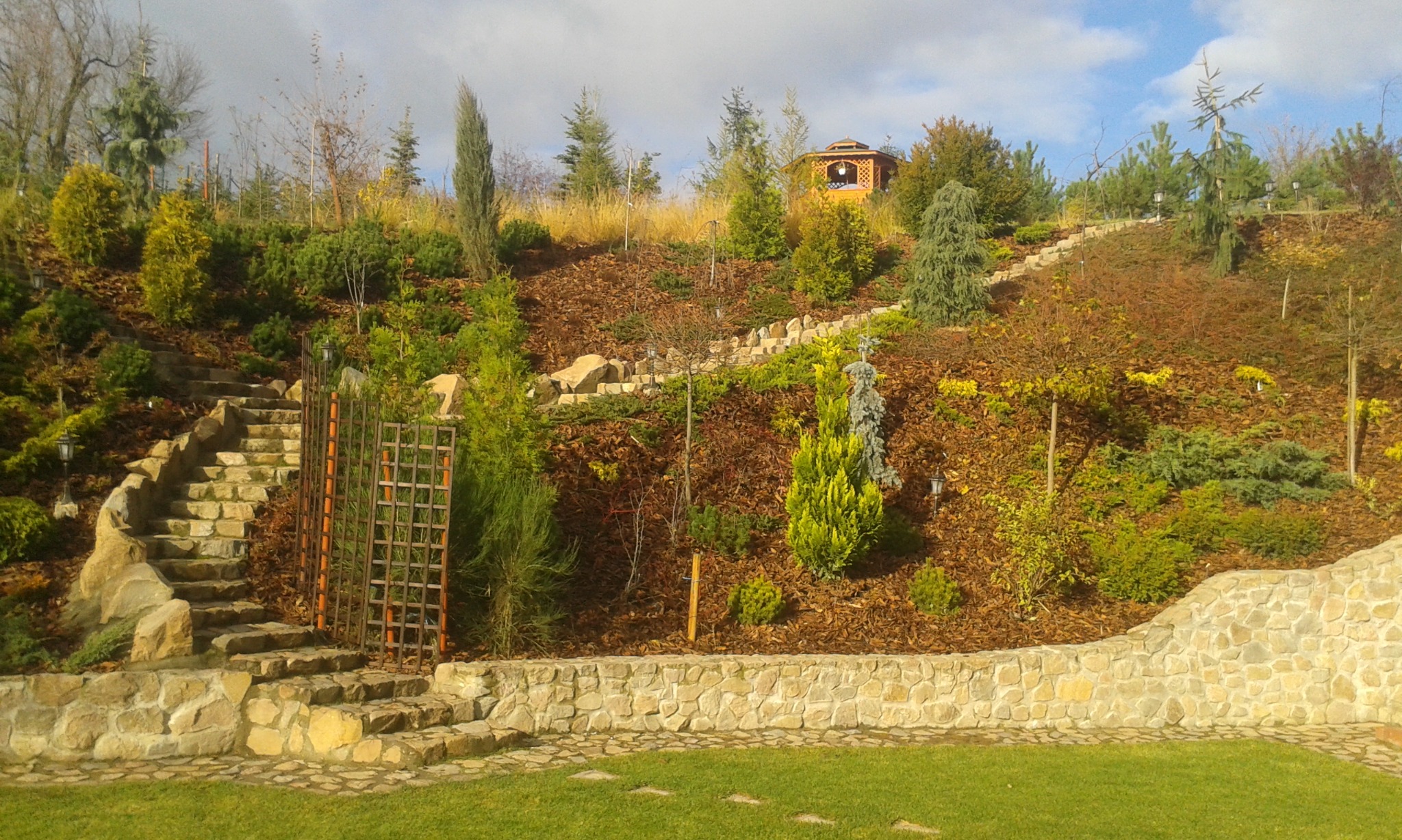 Ukształtowany teren ogrodu ze schodami z kamienia prowadzącymi na górę, gdzie znajduje się drewniana altana, liczne krzewy i drzewa iglaste oraz kamienne murki oporowe.