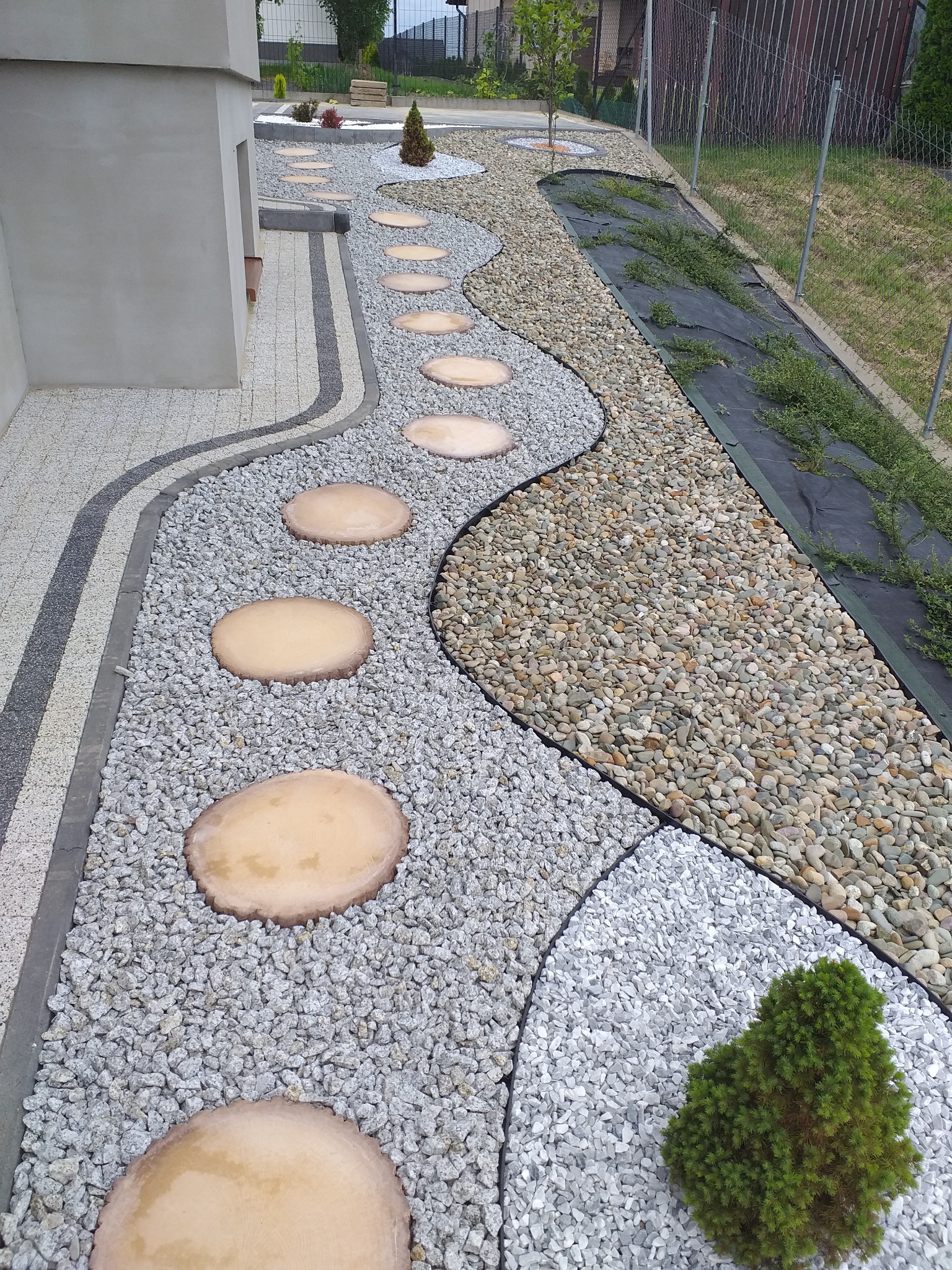 Ogród z ścieżką z okrągłych płyt imitujących drewno, wysypany szarym i beżowym żwirem, obrzeża z kostki brukowej, fragment trawnika z agrowłókniną i nasadzeniami.